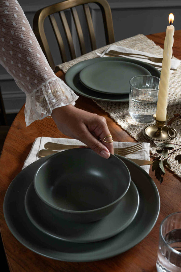 Hand setting a Moss green ceramic dinnerware set from Hamkke Home on a wooden table with brass flatware, glassware, and a lit taper candle.