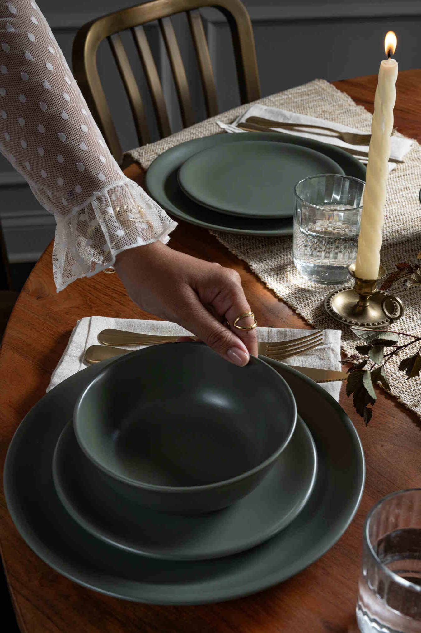 Hand setting a Moss green ceramic dinnerware set from Hamkke Home on a wooden table with brass flatware, glassware, and a lit taper candle.