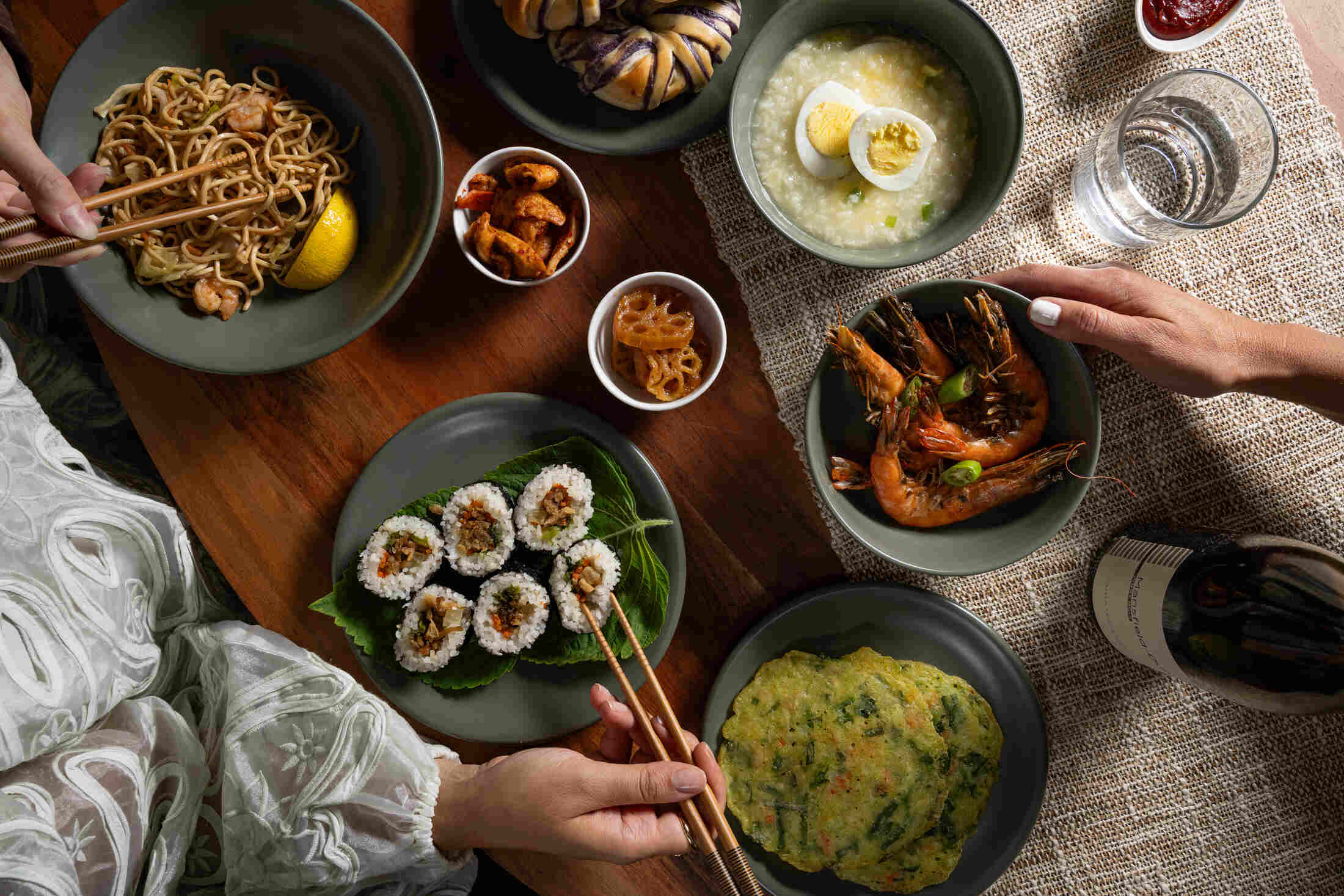 Korean and Asian dishes served on Moss green ceramic plates and bowls from Hamkke Home, including noodles, kimbap, porridge, shrimp, kimchi, and jeon, styled on a wooden table with hands using chopsticks.