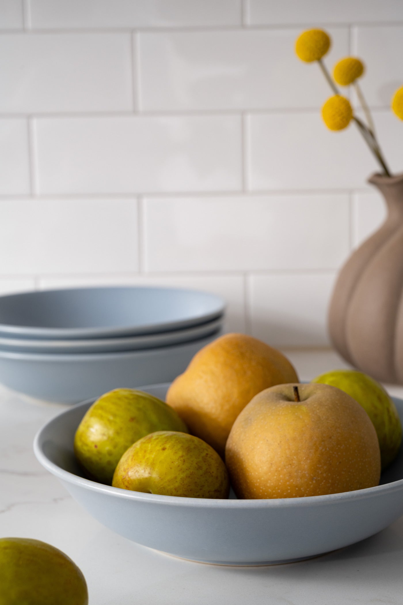 Wave blue ceramic bowls from Hamkke Home filled with Asian pears and figs, styled on a white countertop with a vase of yellow billy balls.