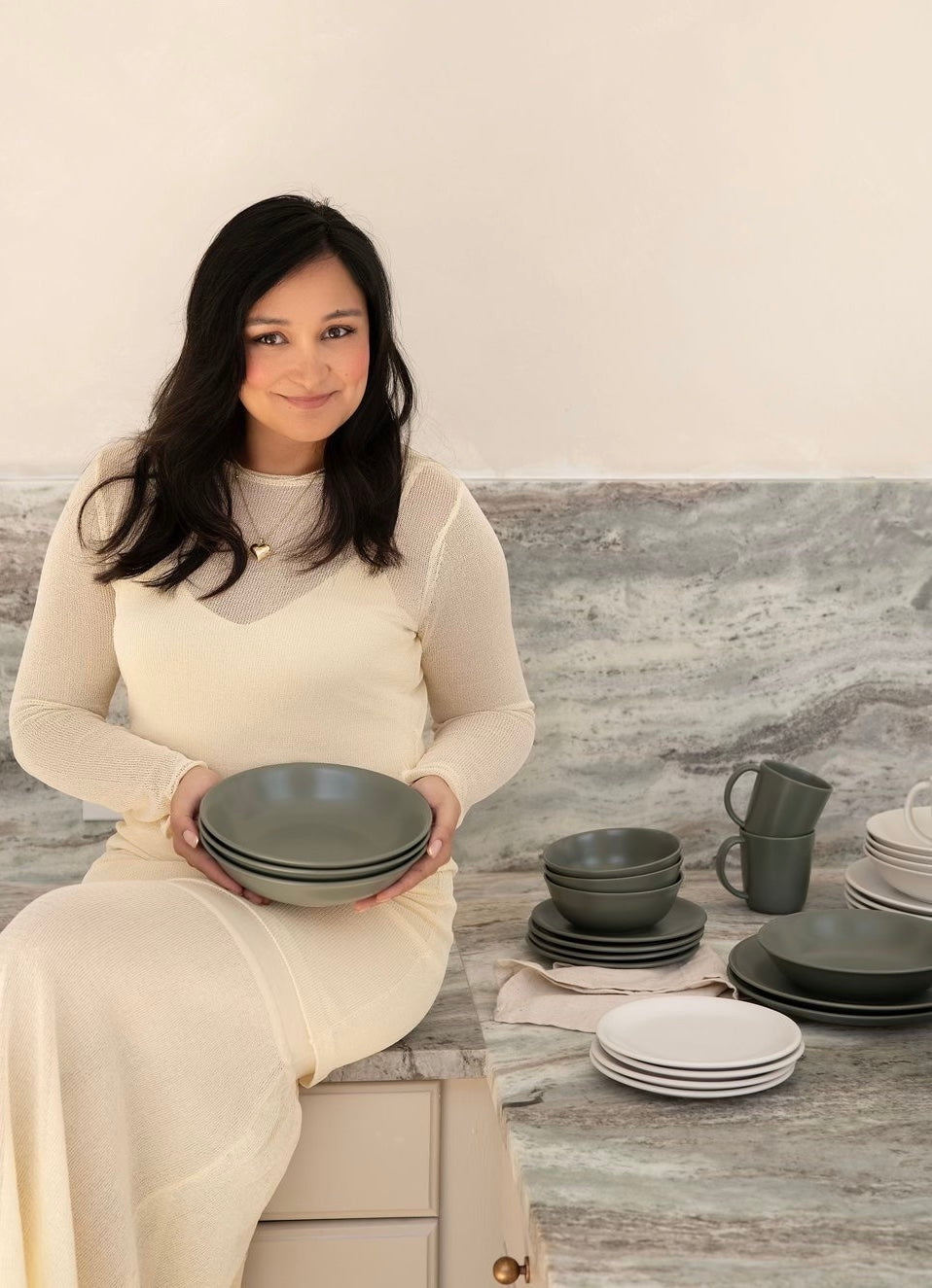 Woman founder seated on a stone countertop holding stacked green ceramic bowls, with matching plates and mugs arranged beside her in a neutral, modern kitchen setting.